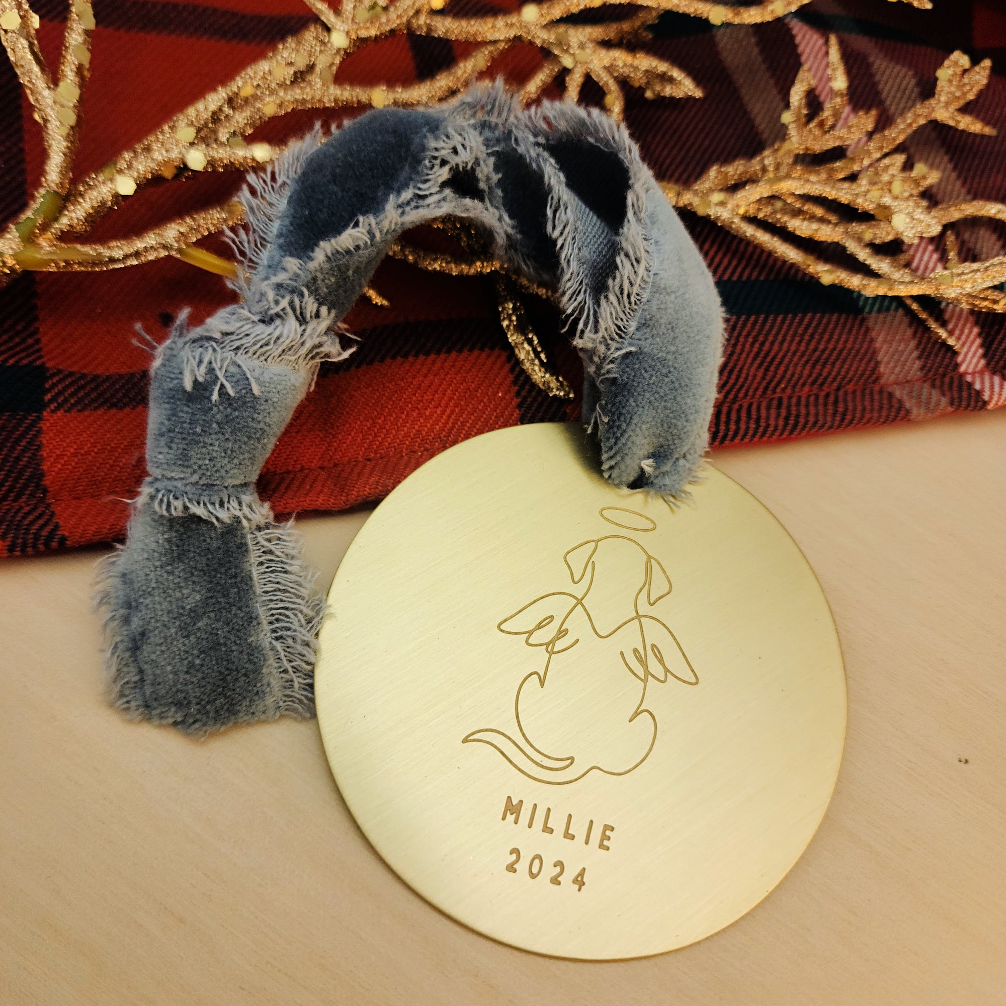 Gold ornament with engraved dog design and 'MILLIE 2024' text, placed on a wooden surface with decorative branches and plaid fabric in the background.