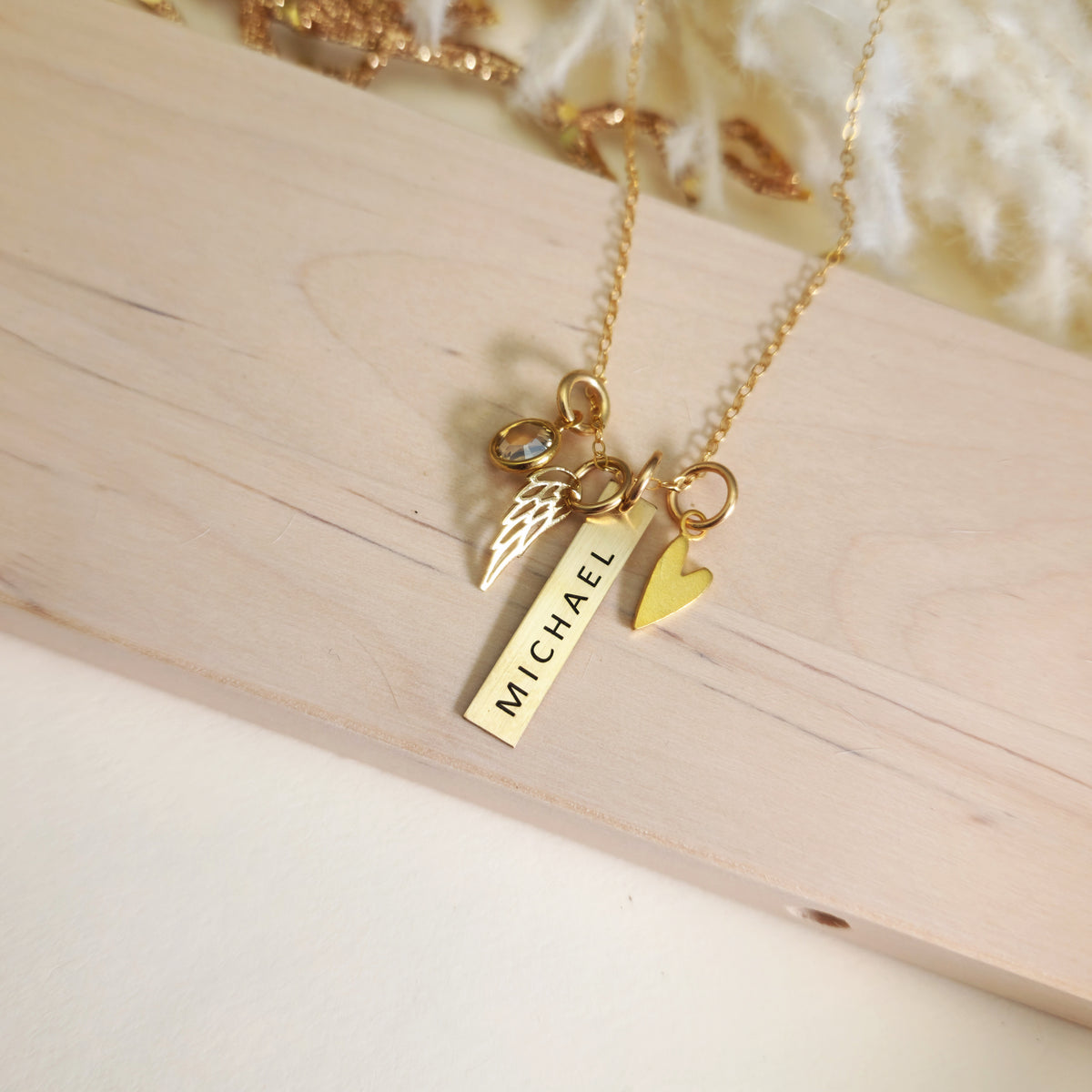 Overhead view of gold memorial necklace with “Michael” engraved bar, heart charm, angel wing charm, and birthstone on wood.