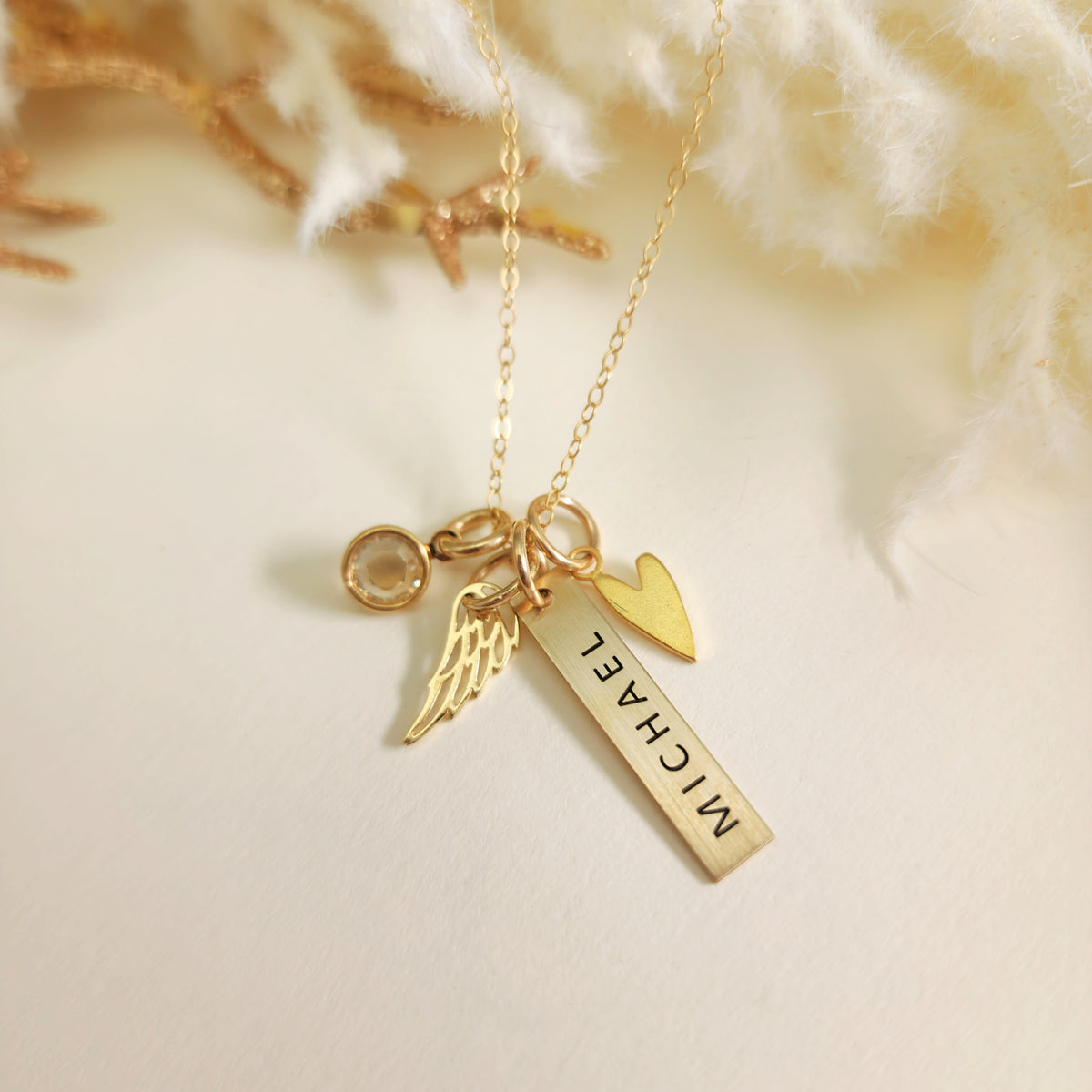 Personalized gold bar memorial necklace with “Michael” engraving, heart charm, angel wing charm, and clear birthstone, on cream background.