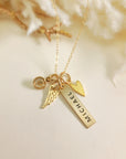 Personalized gold bar memorial necklace with “Michael” engraving, heart charm, angel wing charm, and clear birthstone, on cream background.