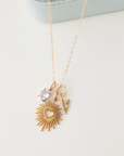 Gold necklace with sunburst pendant and heart-shaped charm on a light gray background