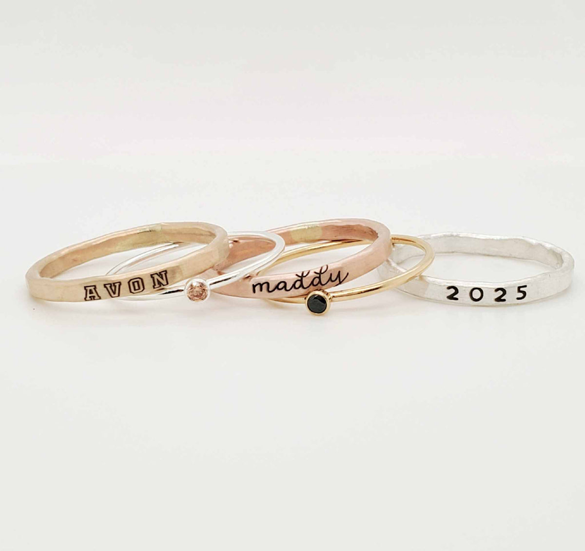 A set of three stackable rings made of yellow gold and sterling silver with two gemstones, and personalized engravings including 'AVON', 'class of 2025', and 'MADDY'.