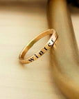 A 2mm gold stacking name ring is resting on a gold bar. The ring has a name engraved in dark font on the center of the ring. There is a visible yellow solder spot on the back of the ring.
