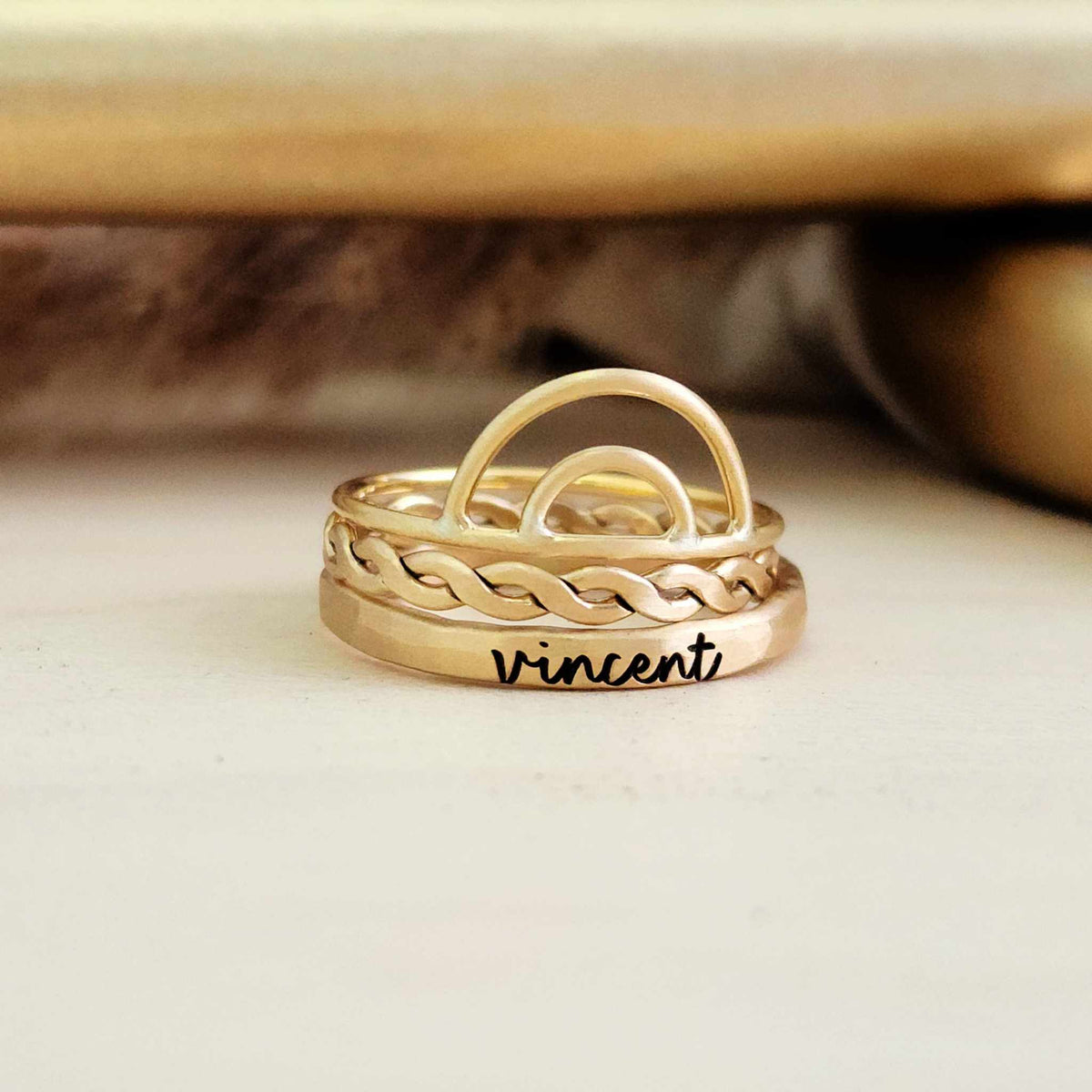 A set of three rings, with the top ring featuring a rainbow design, the middle ring engraved with the name 'Vincent', and the bottom ring with a hammered woven texture. They appear to be made of a gold-filled or sterling silver metal.