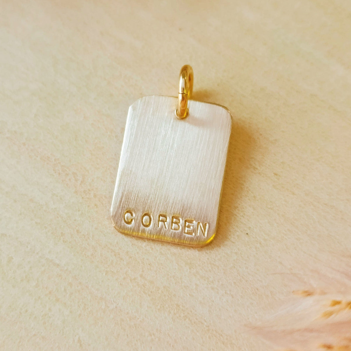 Personalized rectangle charm stamped with the name “CORBEN,” shown without a chain, on a warm-toned wooden background with dried flowers.