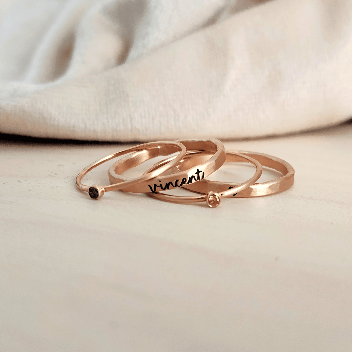 A set of three gold-colored rings, two with engraved names and one with a birthstone, displayed on a person's finger.