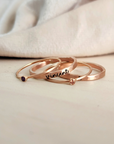 A set of three gold-colored rings, two with engraved names and one with a birthstone, displayed on a person's finger.
