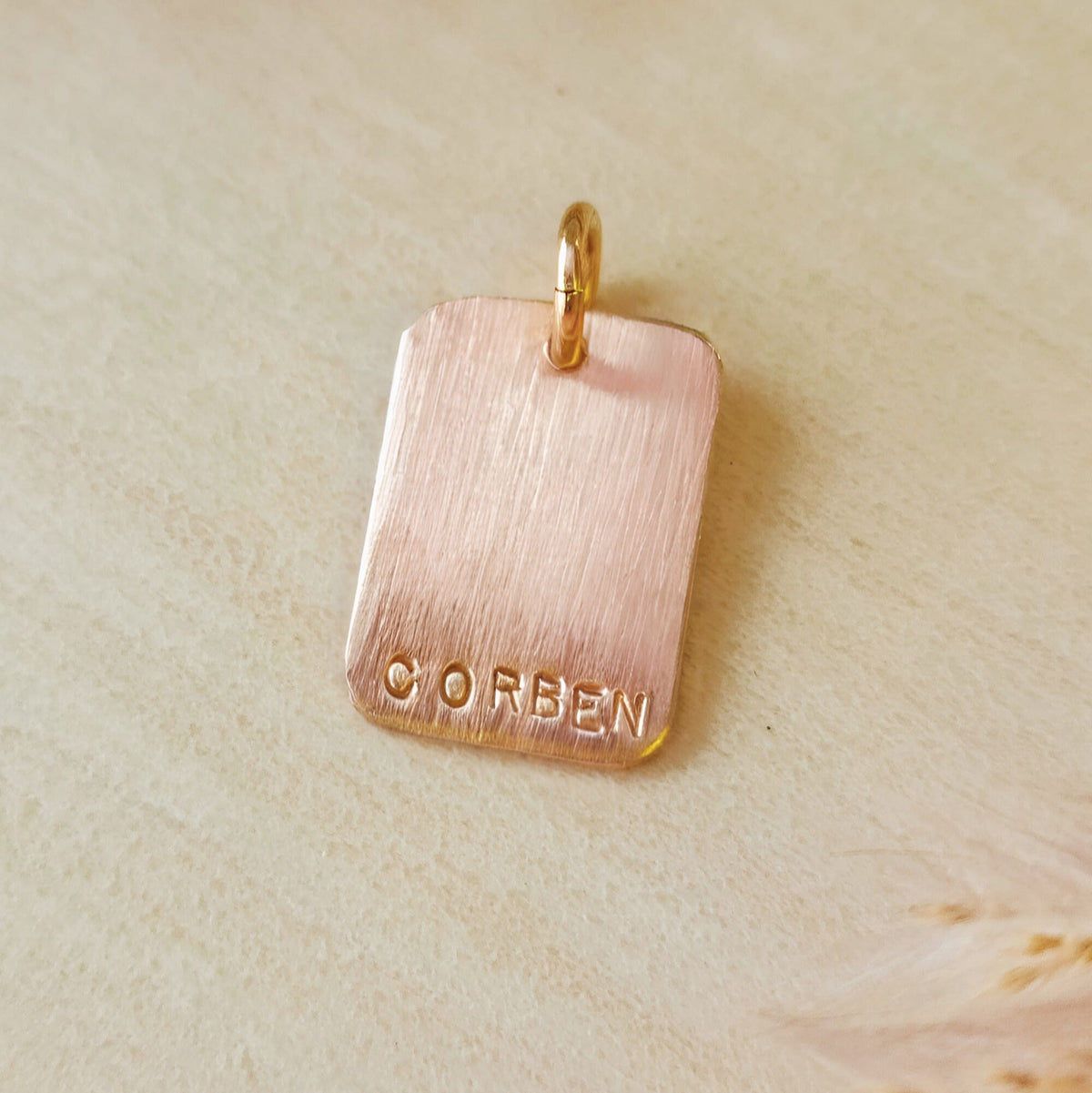 Rose gold pendant with 'CORBEN' engraving on a beige background