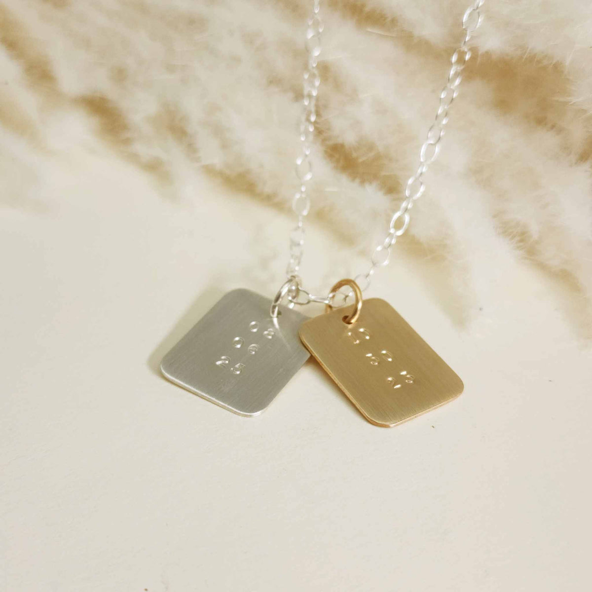 Silver and gold date-stamped pendant necklaces layered together on a silver chain, displayed on a cream background.