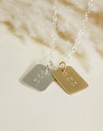 Silver and gold date-stamped pendant necklaces layered together on a silver chain, displayed on a cream background.