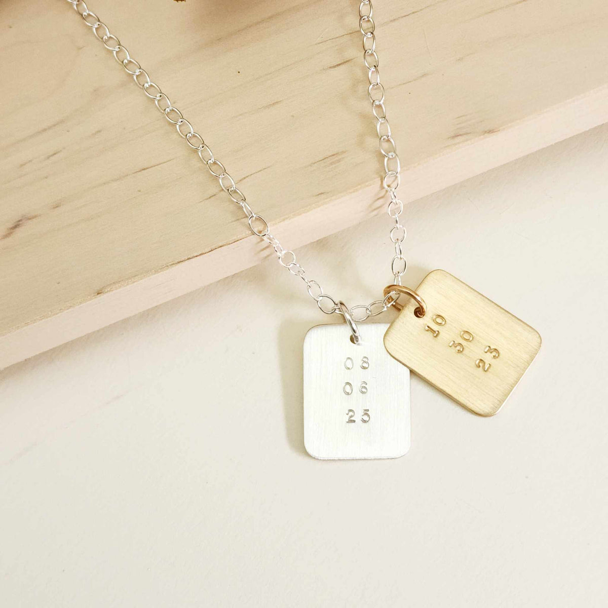 Two rectangular date-stamped pendants, one gold and one silver, on a silver chain displayed on a light wood background.