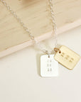 Two rectangular date-stamped pendants, one gold and one silver, on a silver chain displayed on a light wood background.