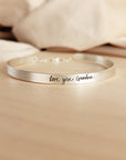 A silver cuff bracelet with custom handwritten engraving, displayed on a wooden surface.