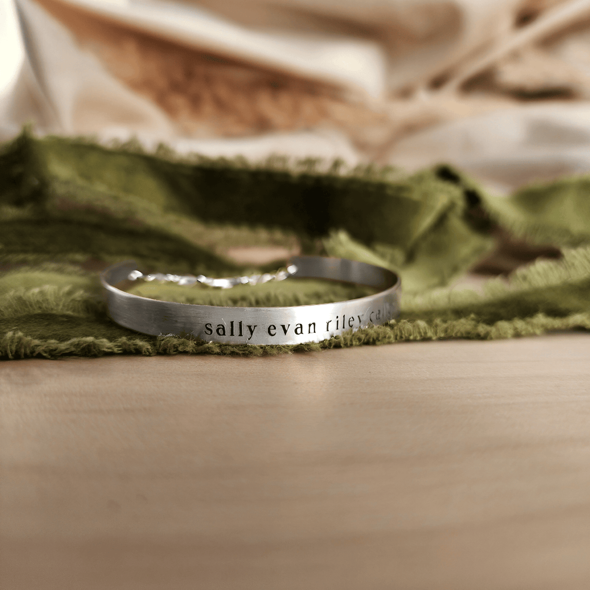 A gold-colored cuff bracelet with custom names engraved on it, displayed on a white surface with a green textured object to the side.