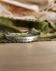 A gold-colored cuff bracelet with custom names engraved on it, displayed on a white surface with a green textured object to the side.