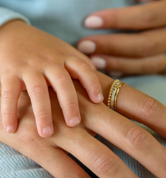 The adult is wearing a set of delicate gold rings, including a beaded band, a thin plain band, and a personalized name ring with an engraved design. The hands are placed against a soft, light blue fabric background, and the adult's nails are neatly manicured with a natural, pale pink polish. The image captures a tender and intimate moment, emphasizing connection and love.