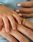 The adult is wearing a set of delicate gold rings, including a beaded band, a thin plain band, and a personalized name ring with an engraved design. The hands are placed against a soft, light blue fabric background, and the adult's nails are neatly manicured with a natural, pale pink polish. The image captures a tender and intimate moment, emphasizing connection and love.