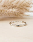 A silver stacking ring with a wavy design, displayed against a neutral background with dry plants in the proximity.