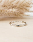 A silver stacking ring with a wavy design, displayed against a neutral background with dry plants in the proximity.
