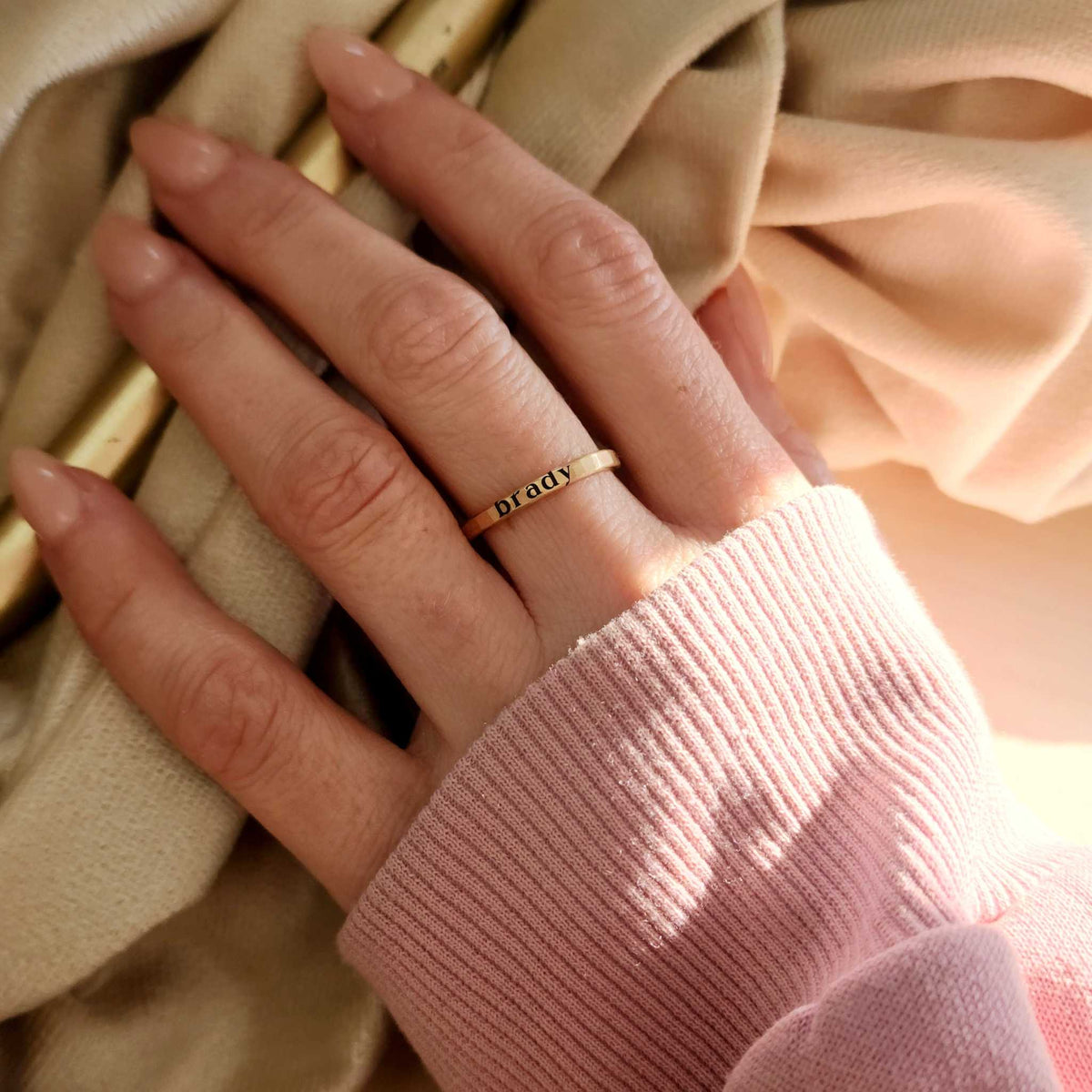 2mm yellow gold stacking ring on the middle finger of a woman with a name engraved on the ring. Woman is wearing a pink sweatshirt with long pink nails.