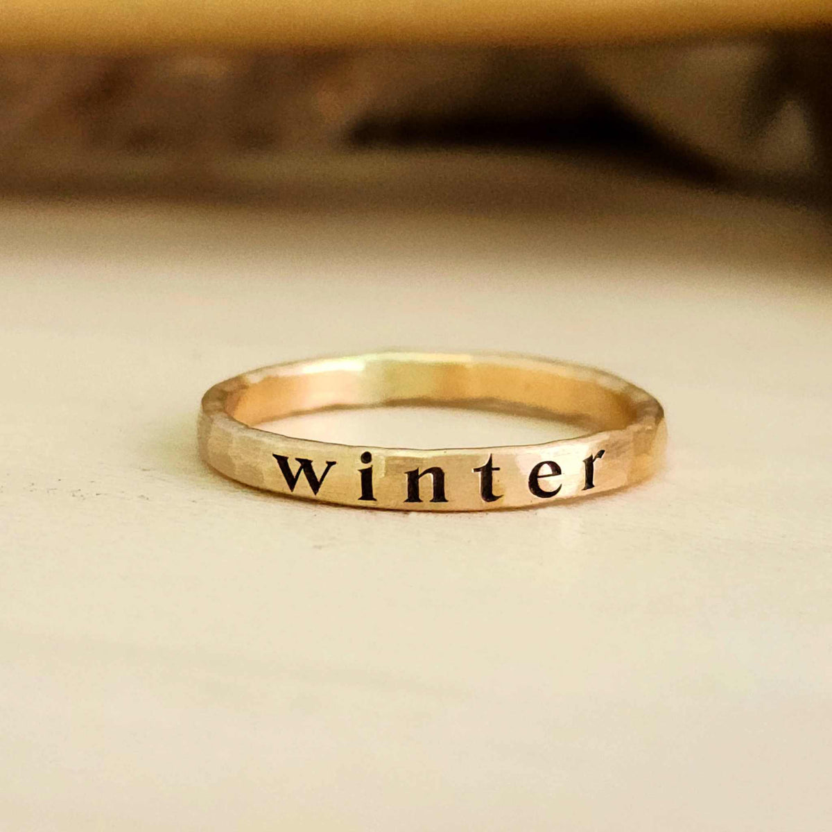 yellow gold ring sitting on a light wood table. Engraved deeply on the ring in serif font, the name "winter". Shadows in background.