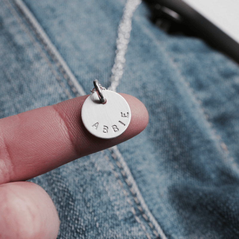 Close-up of a block font name necklace pendant with the name "Abbi" hand stamped on silver metal.