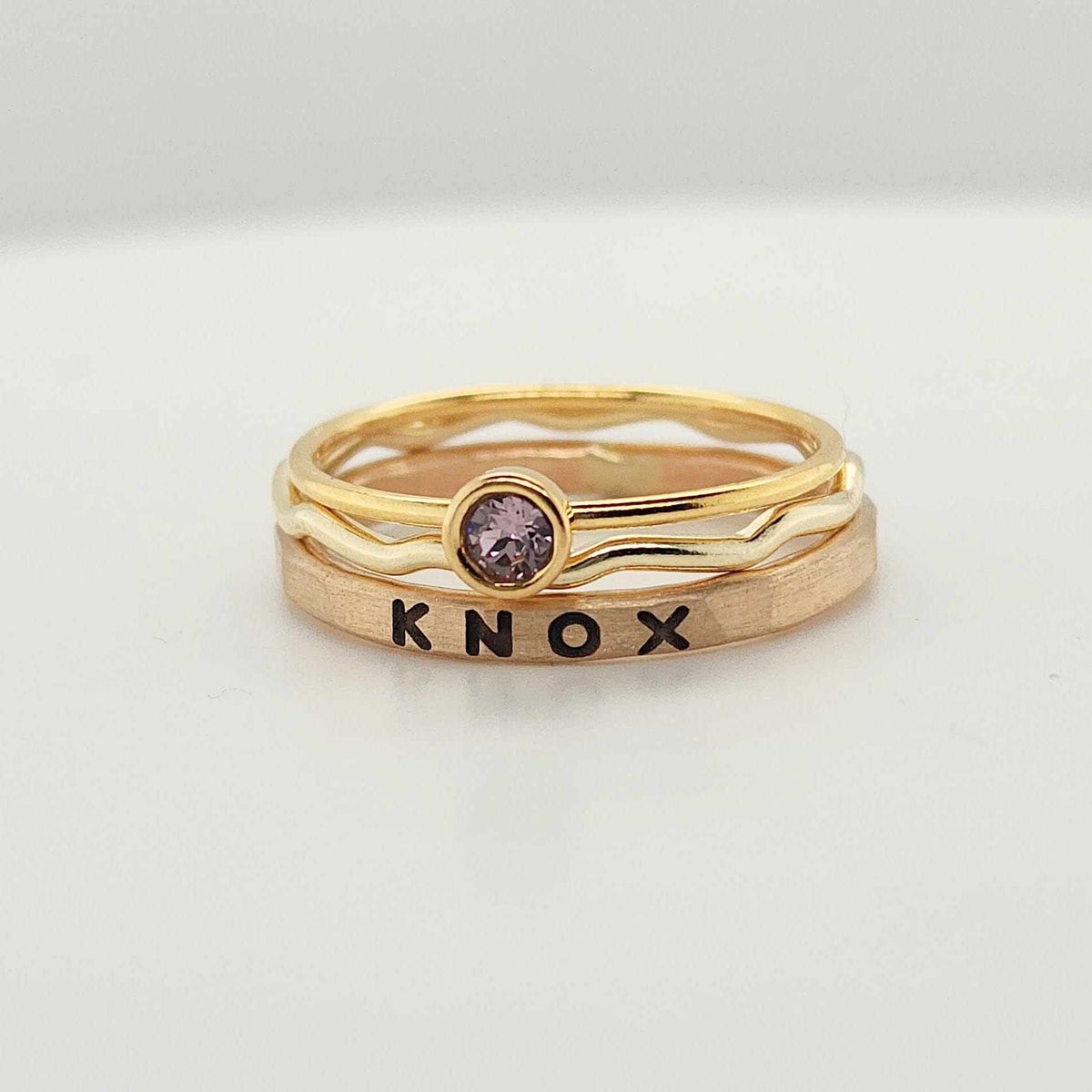 three-piece 14K yellow gold-filled stacking name ring set with personalized name KNOX, wavy ring, and bezel-set birthstone ring