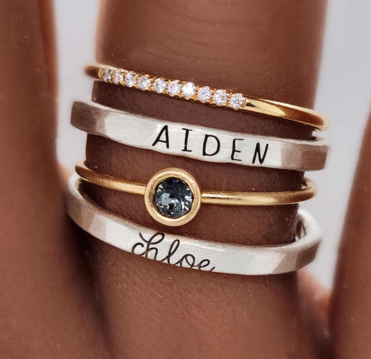 Close-up of a hand wearing the Athena custom name ring set with stacked fine silver and 14K gold-filled rings, personalized with names and a gray gemstone.