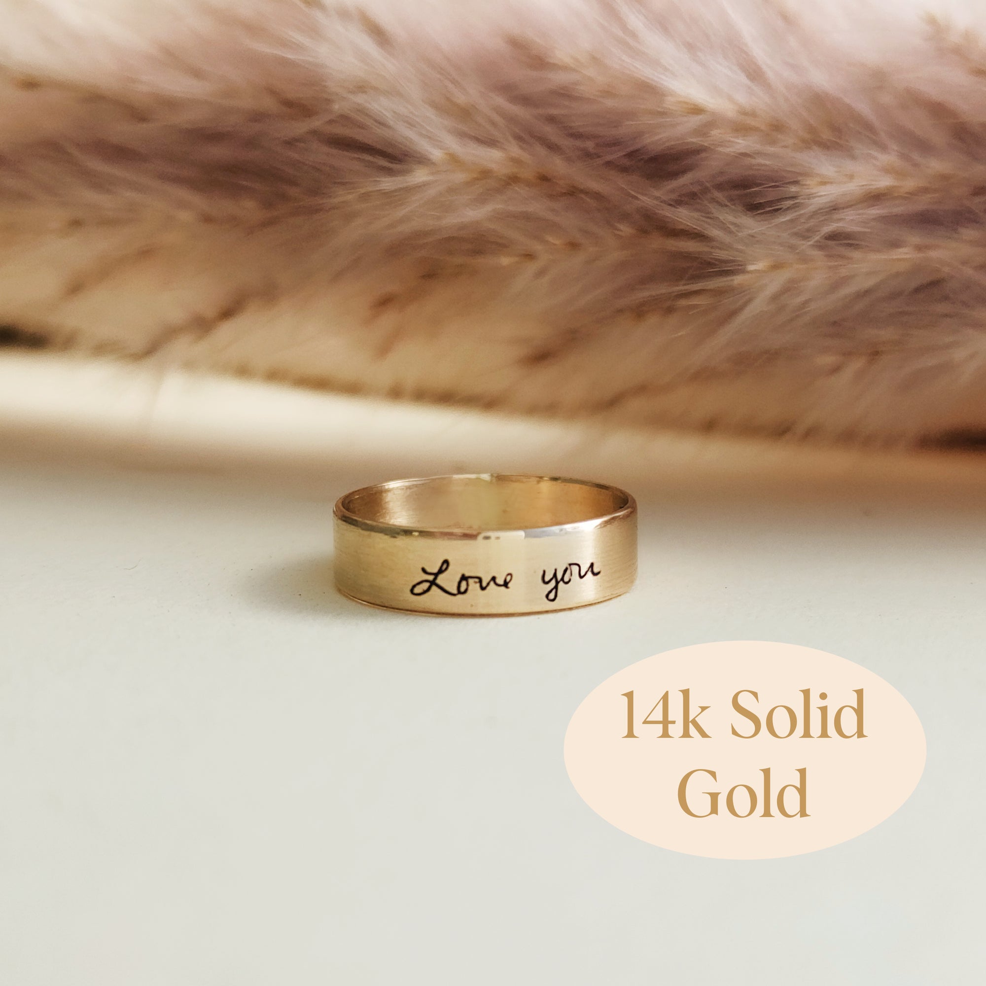 Close-up of 14K solid gold handwriting ring engraved with 'Love you' on soft blush background
