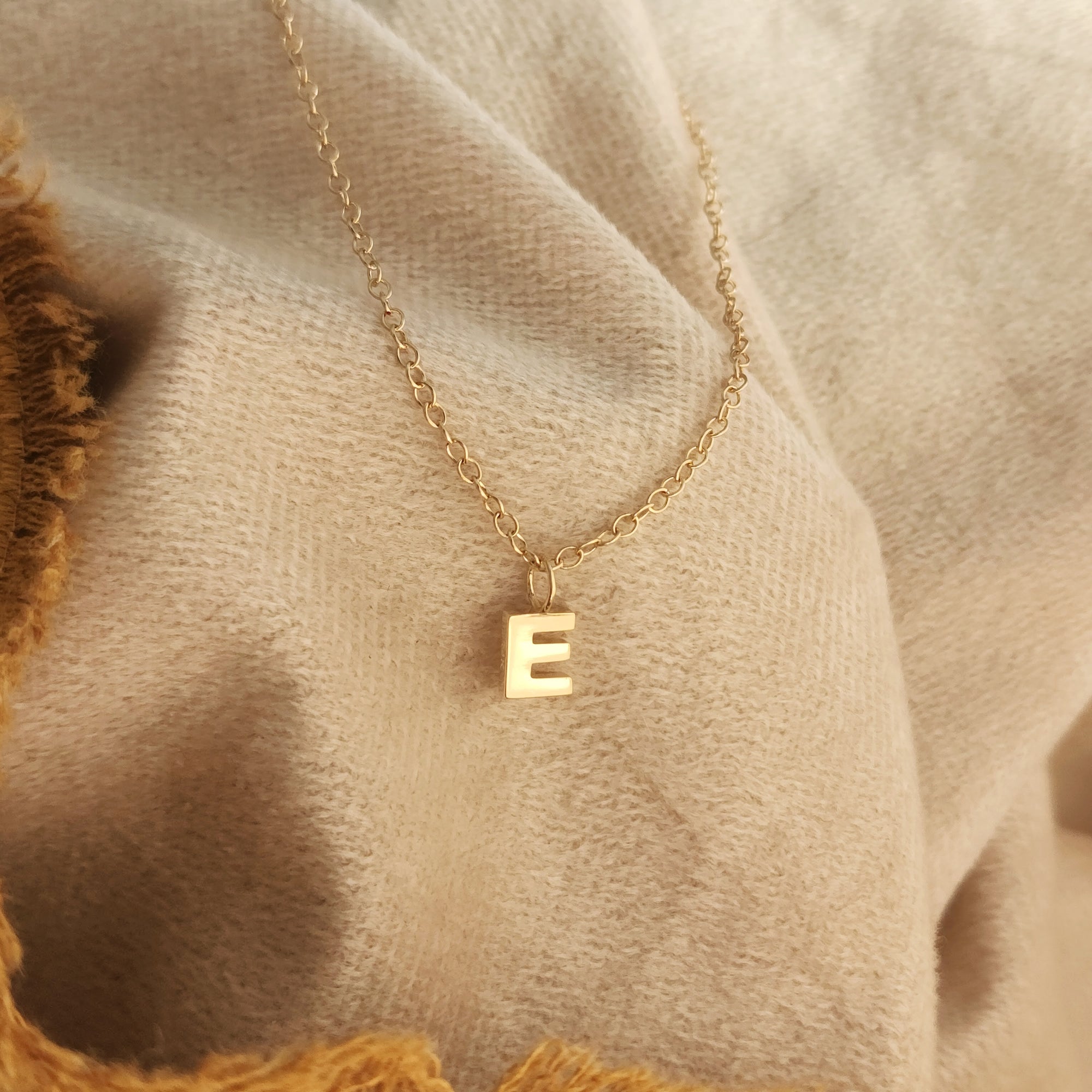 14K solid gold initial pendant necklace with E charm resting on beige linen