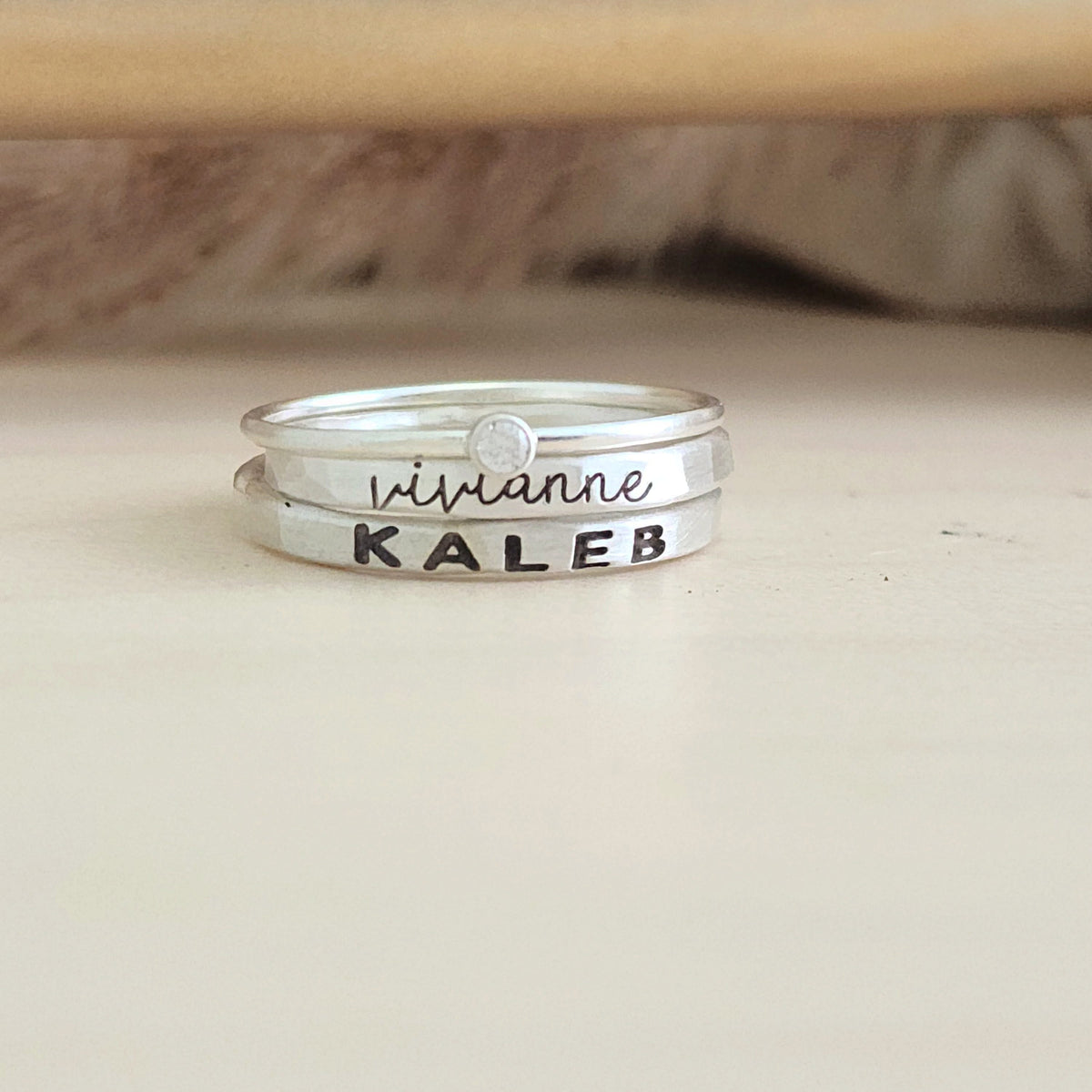 A set of stackable silver rings, with one clear cz ring and two personalized name rings, displayed on a wooden surface.