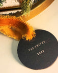 Round black tag with 'The Smiths 2025' text on a decorative background with orange fabric and greenery.