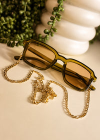 Sunglasses with a gold chain on a light surface with greenery in the background