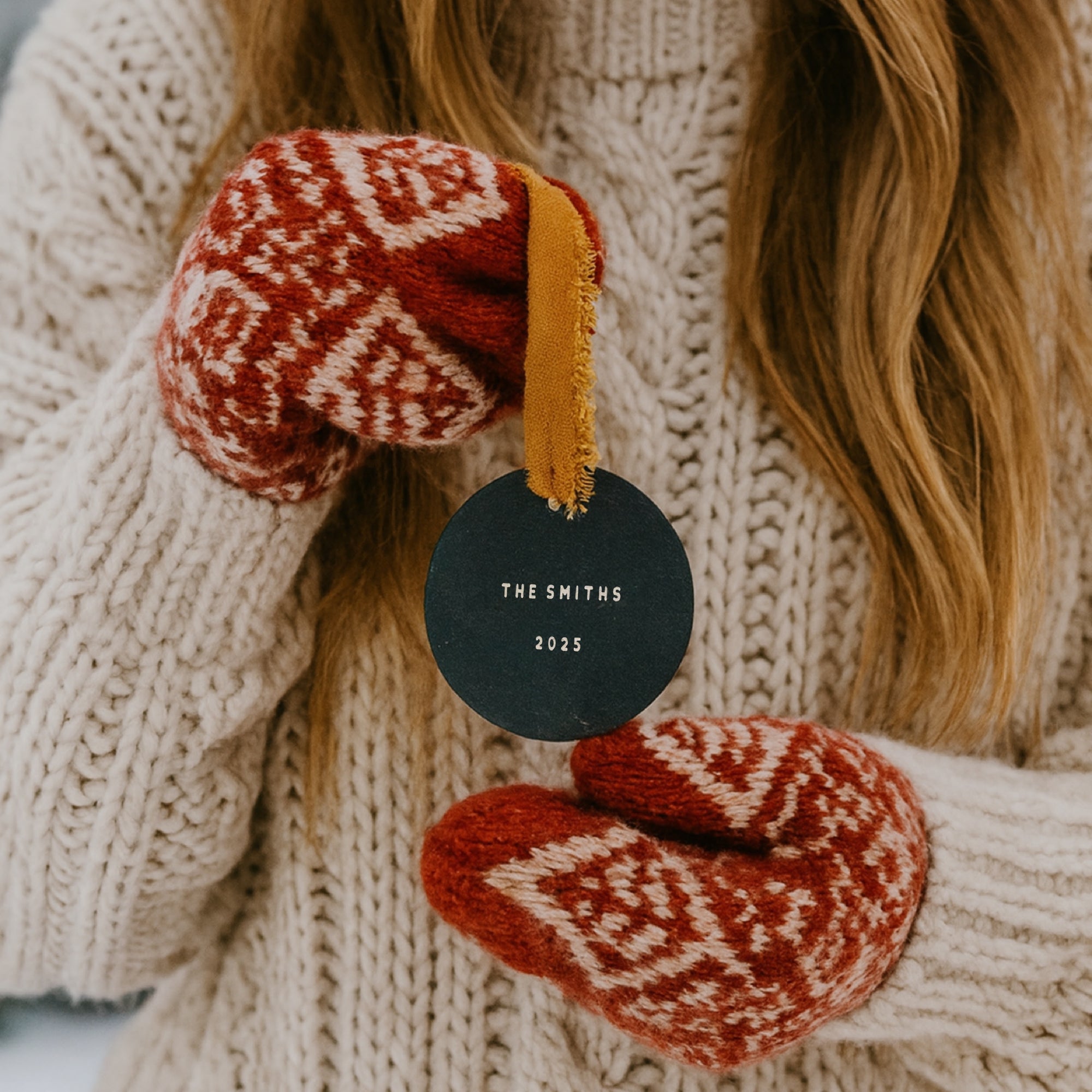 Pair of red and white patterned mittens held by a person wearing a beige sweater, with a black tag reading 'The Smiths 2023'.