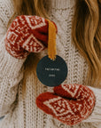 Pair of red and white patterned mittens held by a person wearing a beige sweater, with a black tag reading 'The Smiths 2023'.