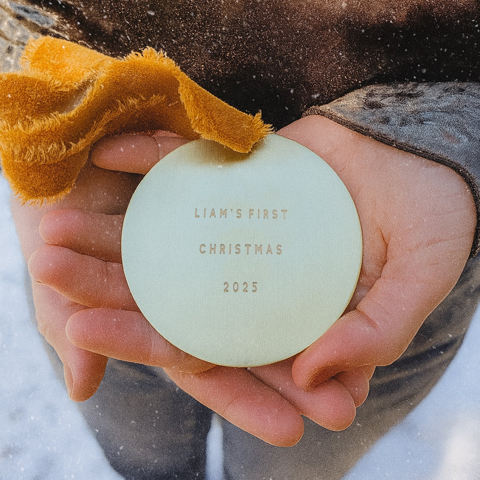 Child's hand holding a light green ornament with 'Liam's First Christmas 2025' text, against a snowy background.