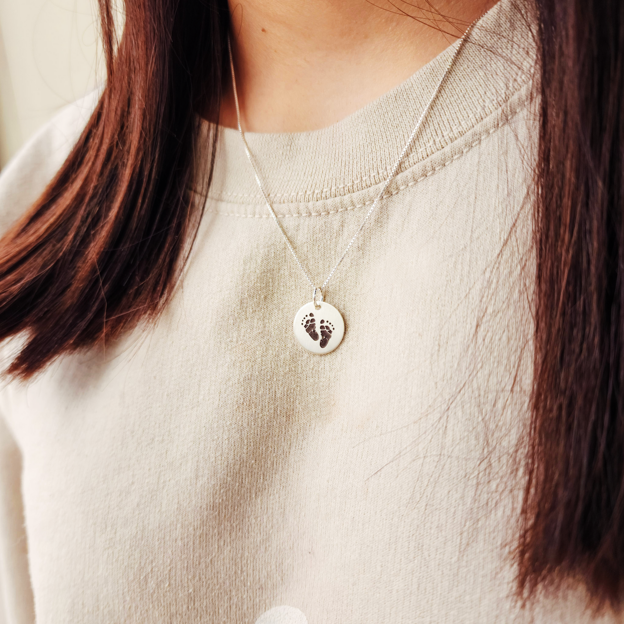 Necklace with a pendant featuring footprints worn by a person in a light-colored sweater.