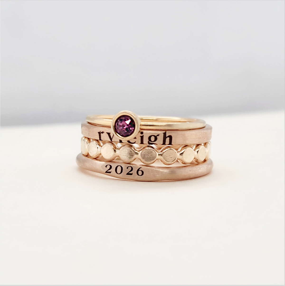 A set of stackable gold rings on a person's hand, featuring personalized engravings such as a name and the year 2026.