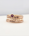 A set of stackable gold rings on a person's hand, featuring personalized engravings such as a name and the year 2026.