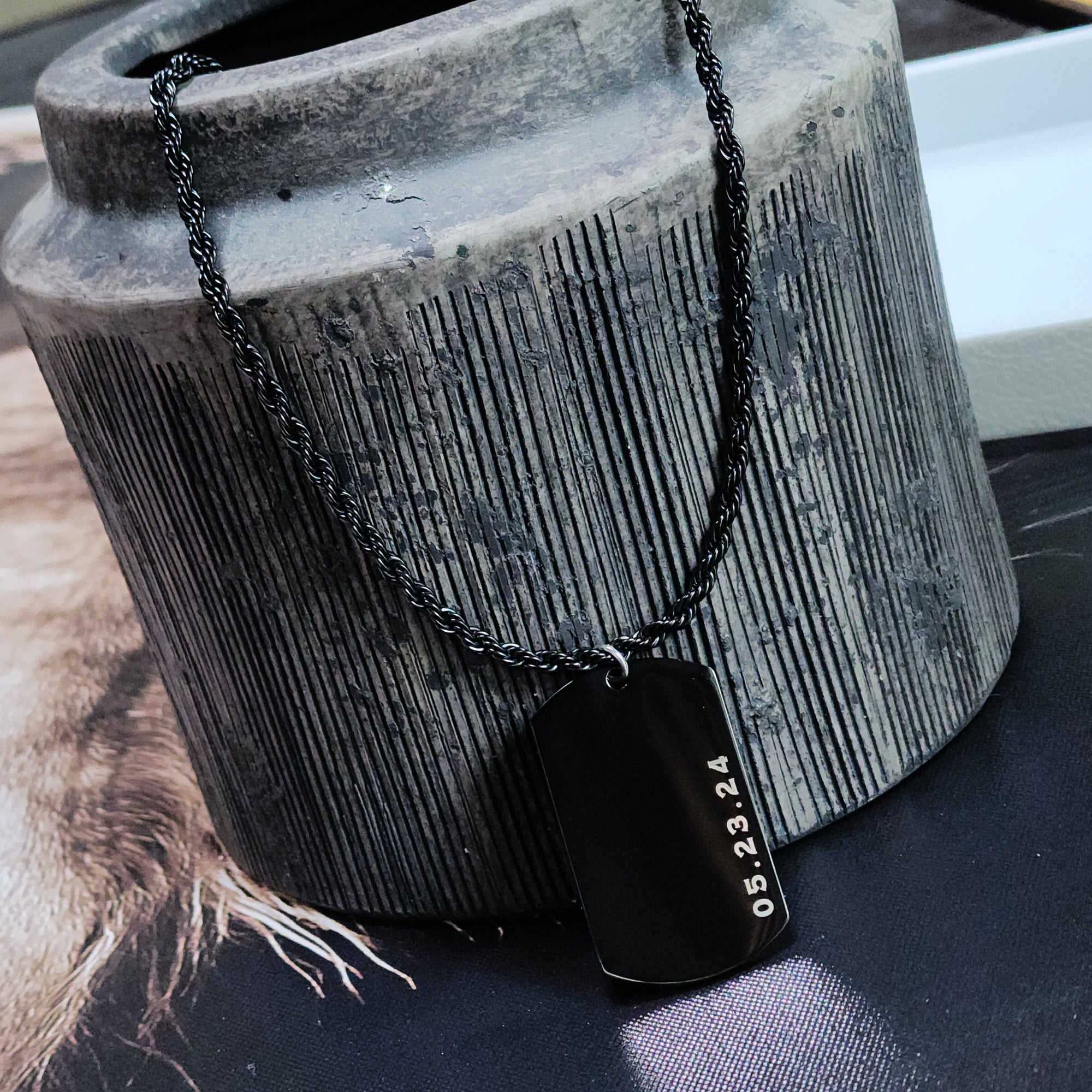 Black dog tag necklace with a date on it, hanging from a textured gray vase.