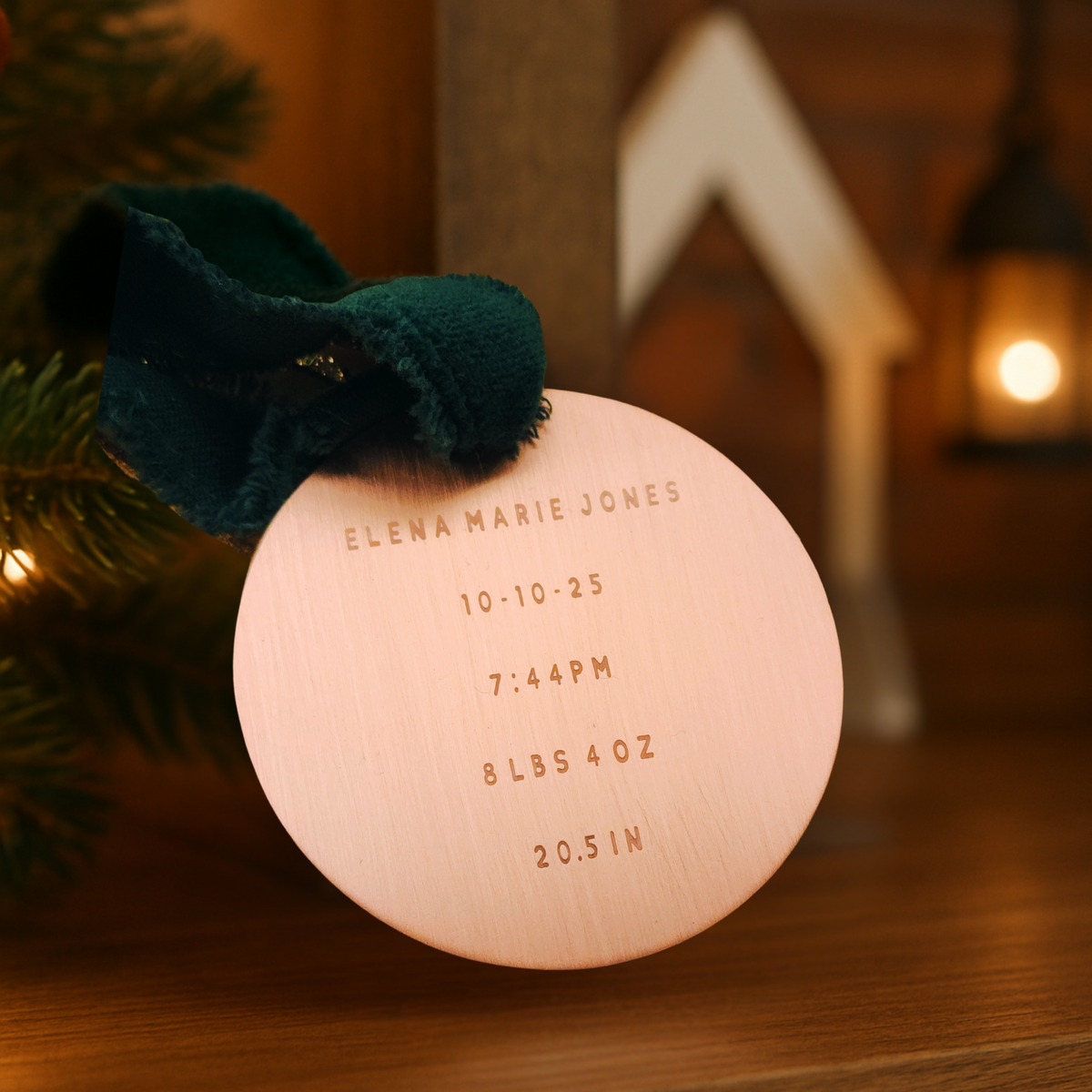 Copper ornament with engraved birth details on a wooden surface with a blurred Christmas tree and lights in the background.