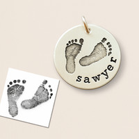 Round silver pendant with footprints and 'sawyer' engraving on a beige background