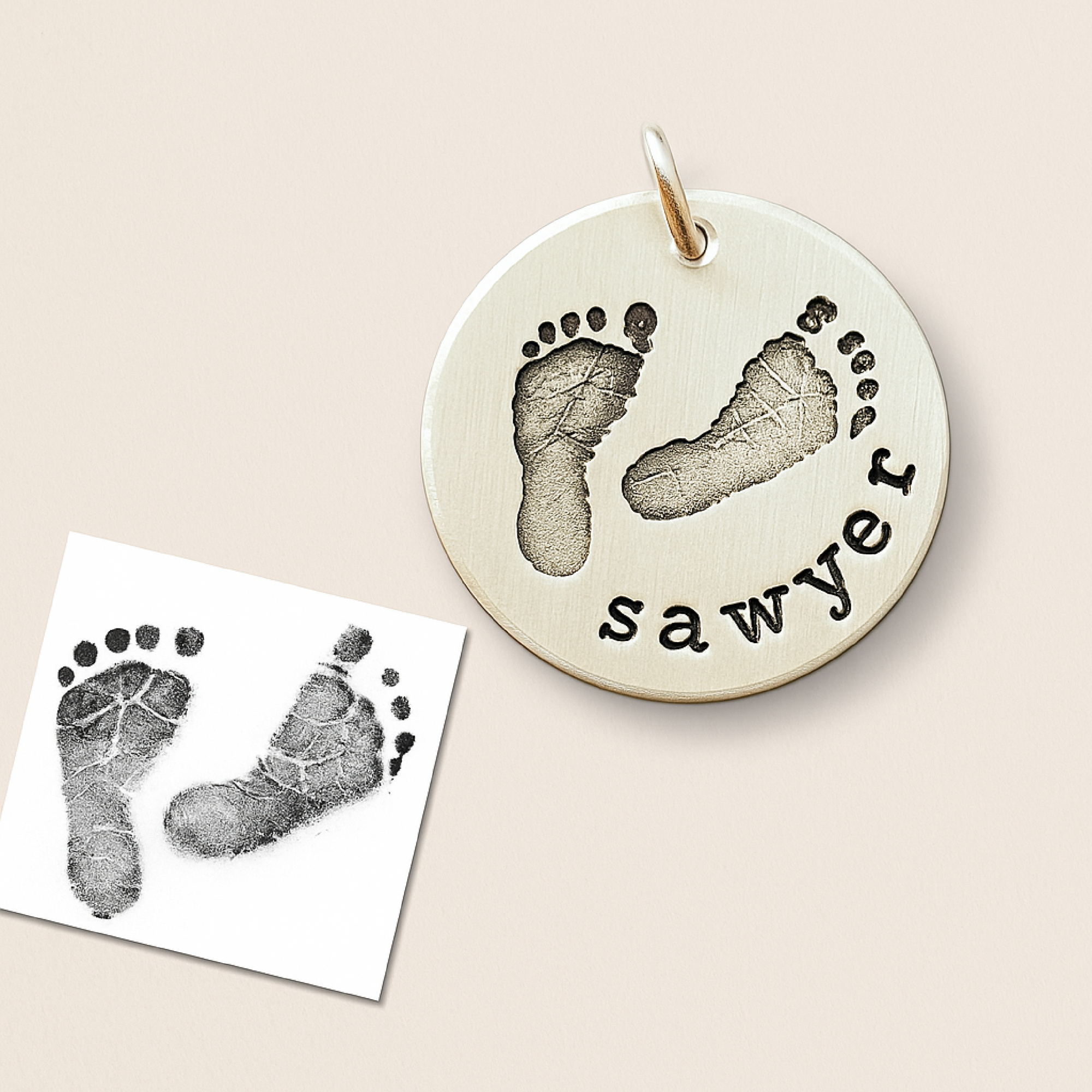 Round silver pendant with footprints and 'sawyer' engraving on a beige background