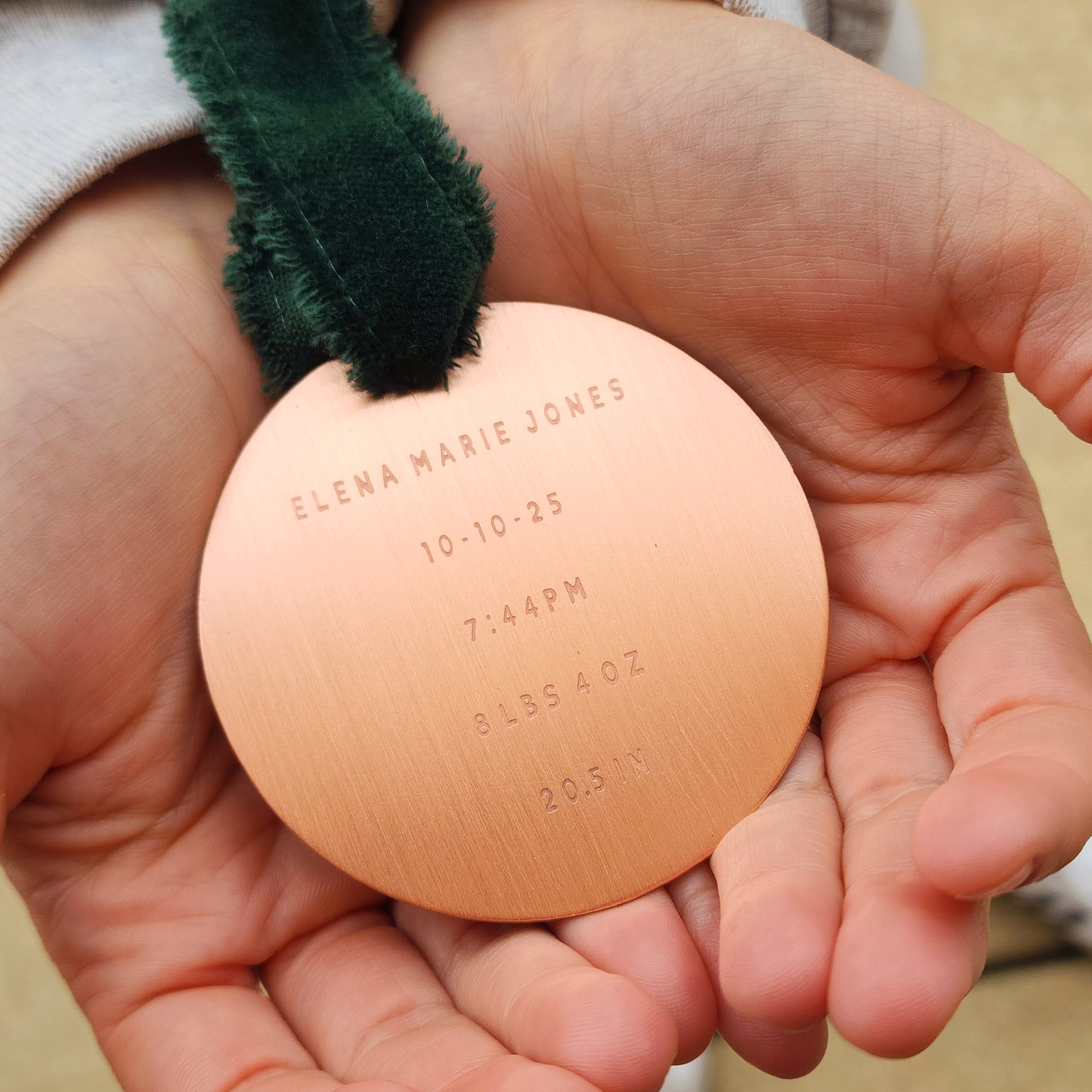 Round copper baby birth announcement ornament held in a hand with green ribbon.