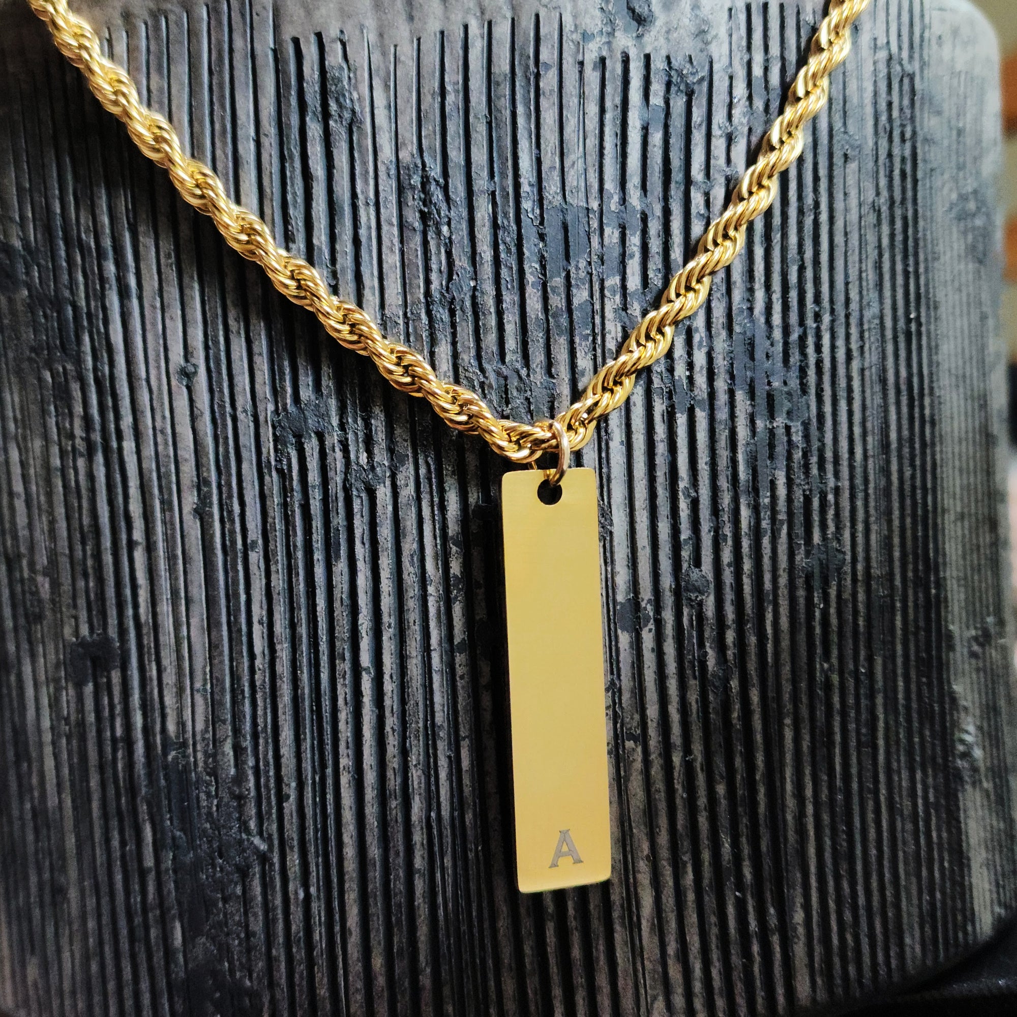 Gold necklace with a vertical bar pendant featuring the letter 'A' on a textured dark surface.