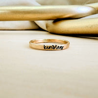 A gold-colored personalized stacking name ring with cursive writing on a textured band, placed on a beige surface with a reflective object in the background.