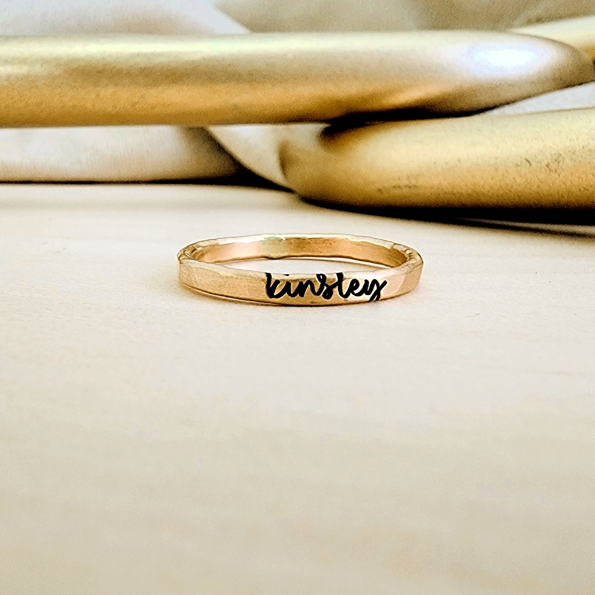 A gold-colored personalized stacking name ring with cursive writing on a textured band, placed on a beige surface with a reflective object in the background.