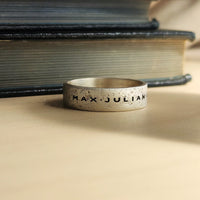 Silver ring with 'MAX JULIAN' engraving on a textured surface with books in the background