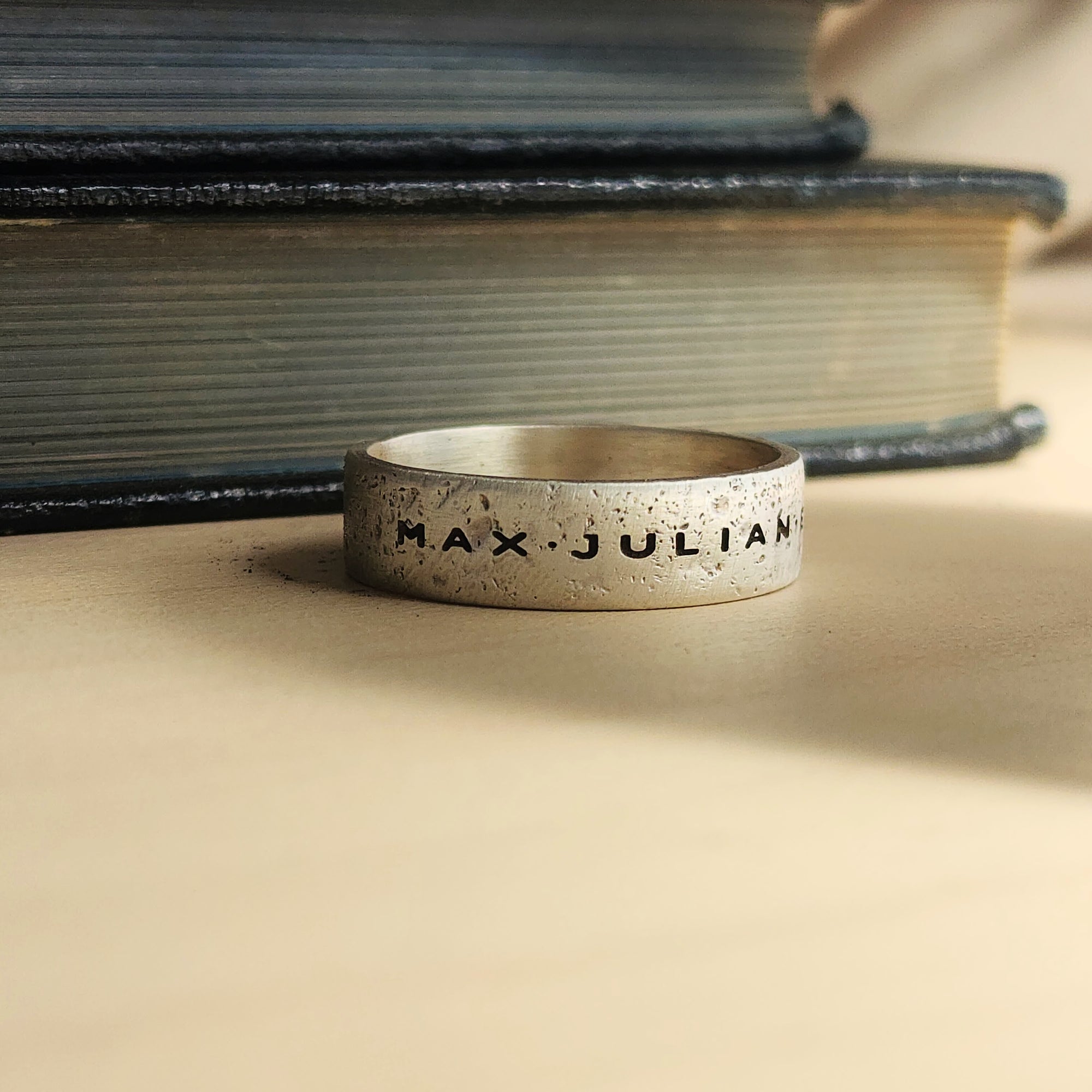 Silver ring with 'MAX JULIAN' engraving on a textured surface with books in the background