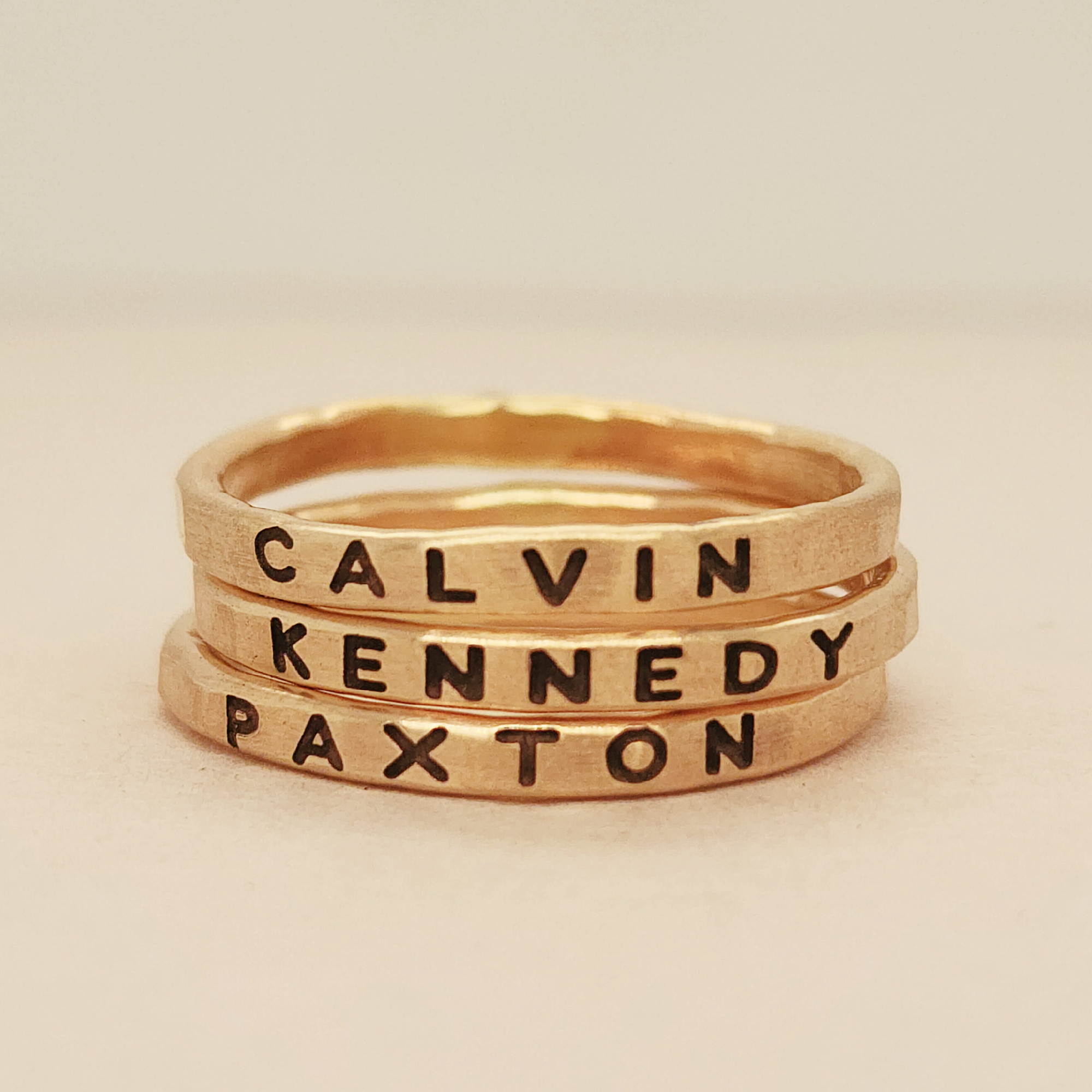 Three gold-colored stacking rings with the names CALVIN, KENNEDY, and PAXTON engraved in bold uppercase letters.